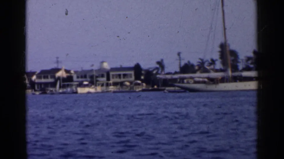 1955 California harbor view with a long sailboat near shoreline houses, palm trees, and low docks under a hazy sky.
