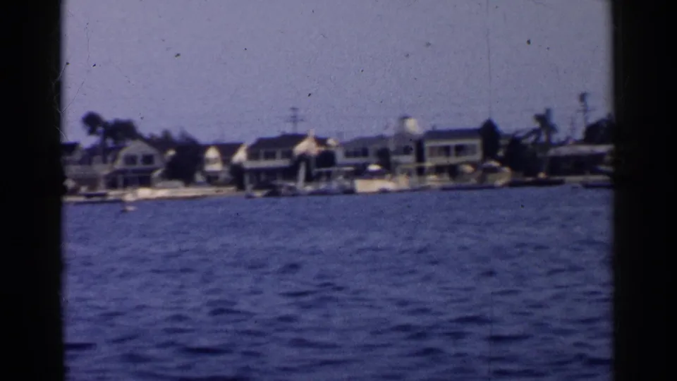 1955 California 8mm frame with shoreline residences, small boats, palms, and rippled harbor water during a waterfront pan.