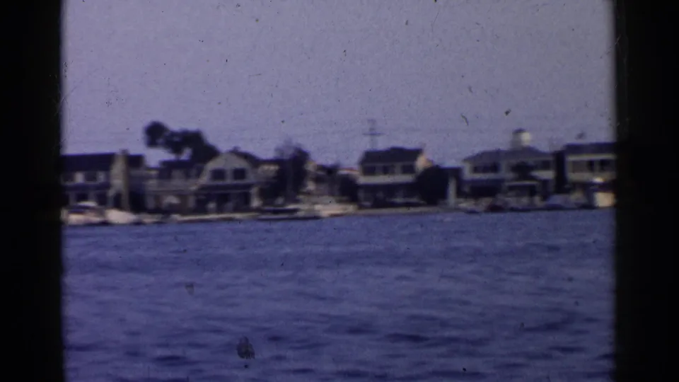 1955 California coastal harbor frame showing waterfront homes, moored boats, palm trees, and blue water within dark 8mm borders.