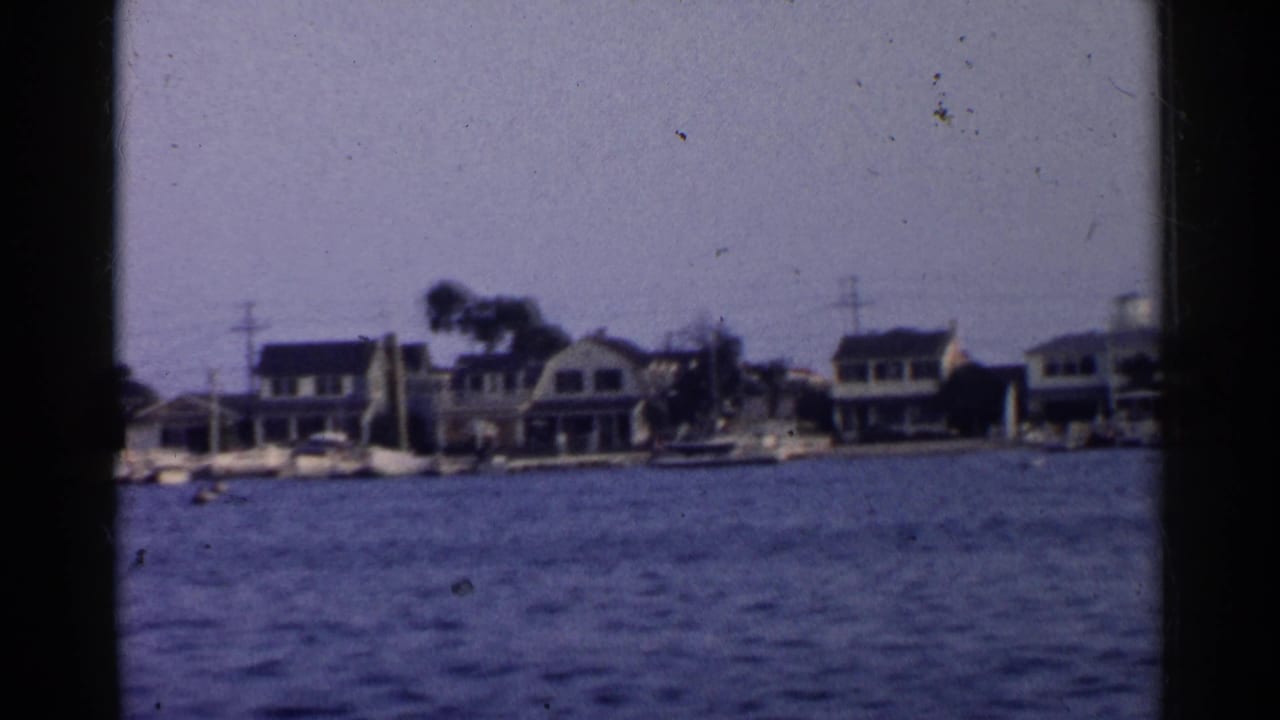 Waterfront houses and docked sailboats seen across rippled water from a dark-framed moving boat view.