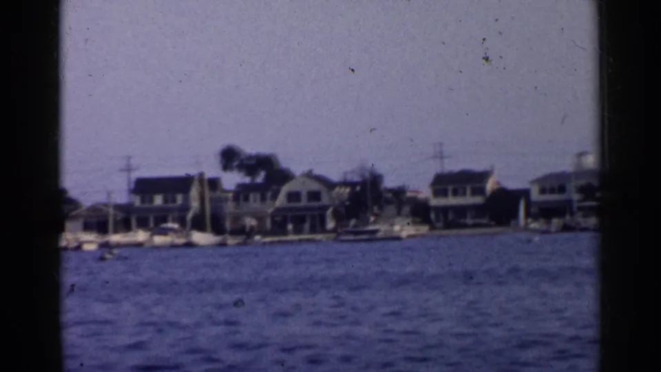 1955 California 8mm frame showing shoreline houses, sailboats, and calm water, partially bordered by a dark boat-window or film edge.