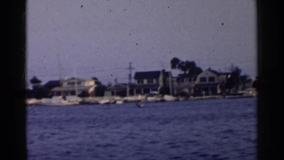 1955 California waterfront view from a moving boat, with residential houses, sailboats, trees, and utility poles along the shore.