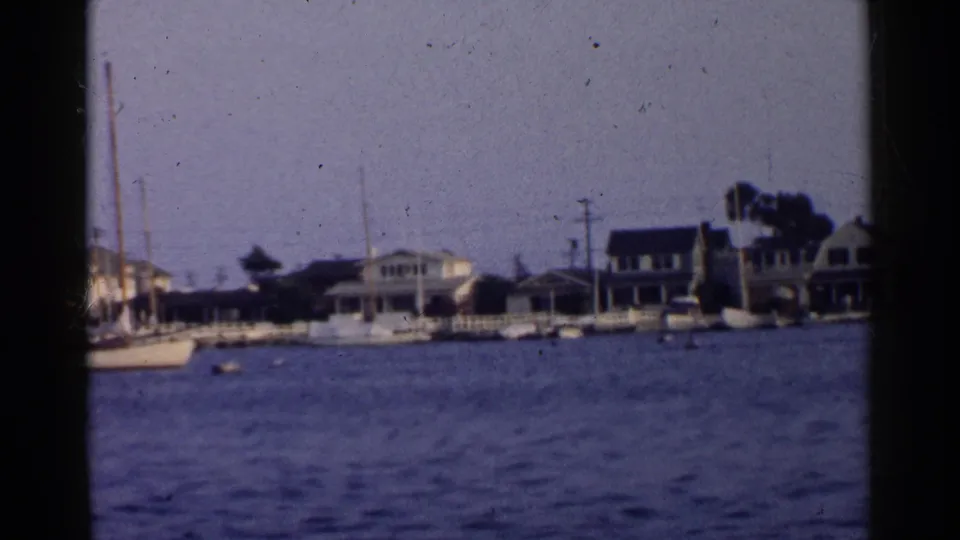Soft 1955 California 8mm frame of a waterfront neighborhood, with sailboats, mooring buoys, docks, and houses along the shoreline.