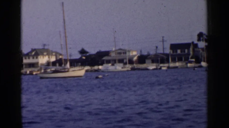 1955 California waterfront frame with a prominent sailboat, docked boats, shoreline homes, and dark film or boat-window borders.