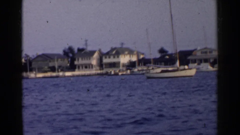 1955 California waterfront scene with low houses, docks, and a sailboat along a residential shoreline.