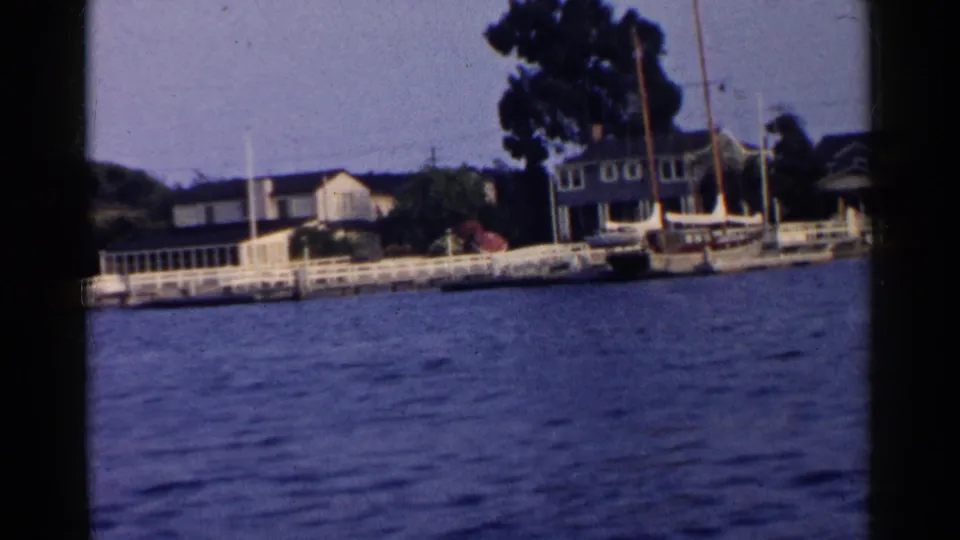 1955 California 8mm frame showing docks, tied boats, trees, and waterfront homes across a rippling waterway.