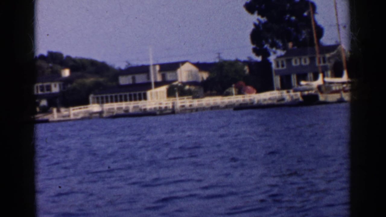 1955 California 8mm frame showing waterfront homes, white railings, shoreline trees, and blue water from a moving boat perspective.
