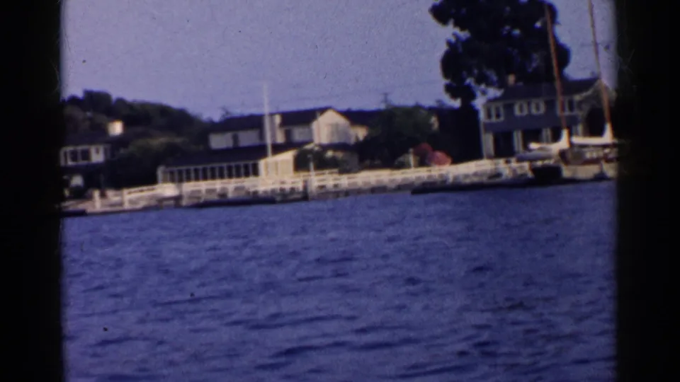 1955 California 8mm frame showing waterfront homes, white railings, shoreline trees, and blue water from a moving boat perspective.