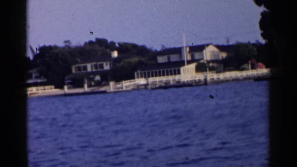 Boat-level 8mm view of California waterfront houses, docks, white railings, and trees along a calm shoreline.