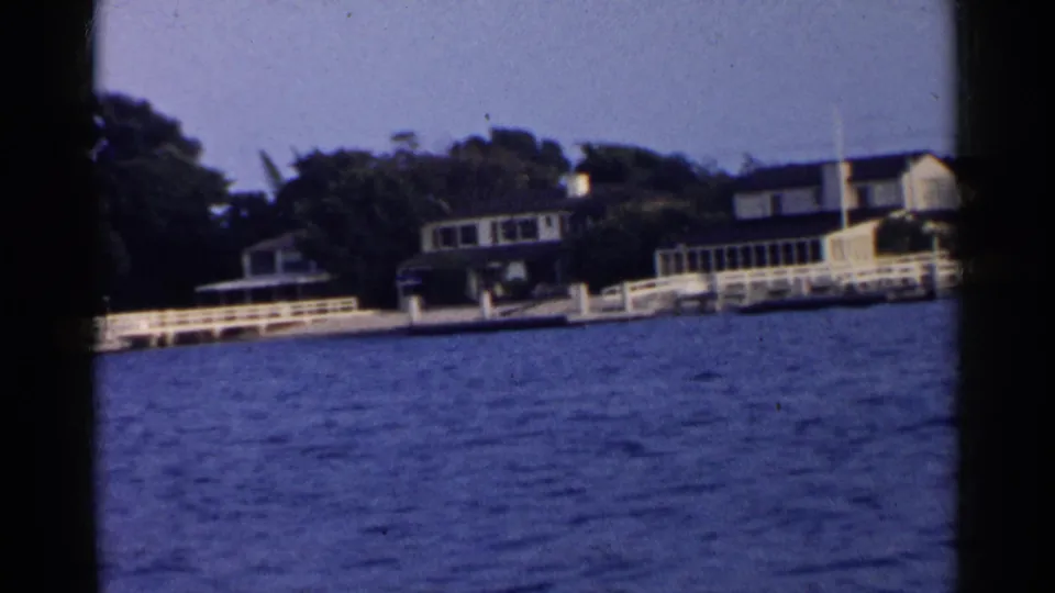 1955 home-movie frame of a waterfront residence, long white railing, trees, and rippled water in California.
