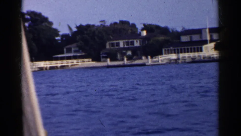 Final 8mm frame showing California waterfront homes behind white railings, with blue water and a pale boat edge in the foreground.