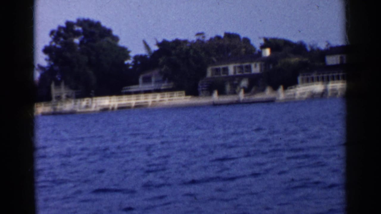 Blue water passing a residential shoreline with houses, trees, railings, and a small moored boat.