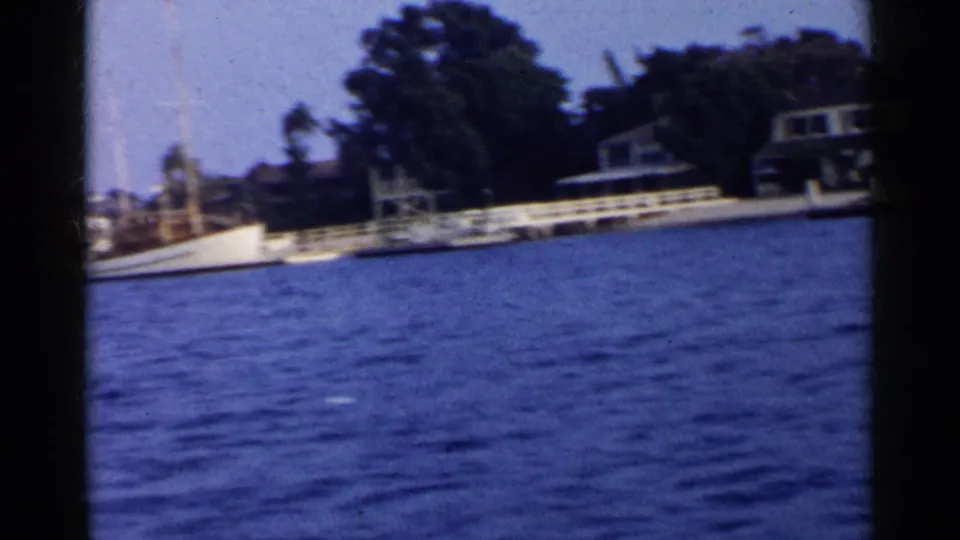 1955 California waterfront view with blue water, shoreline homes, trees, docks, and a moored vessel partly visible.