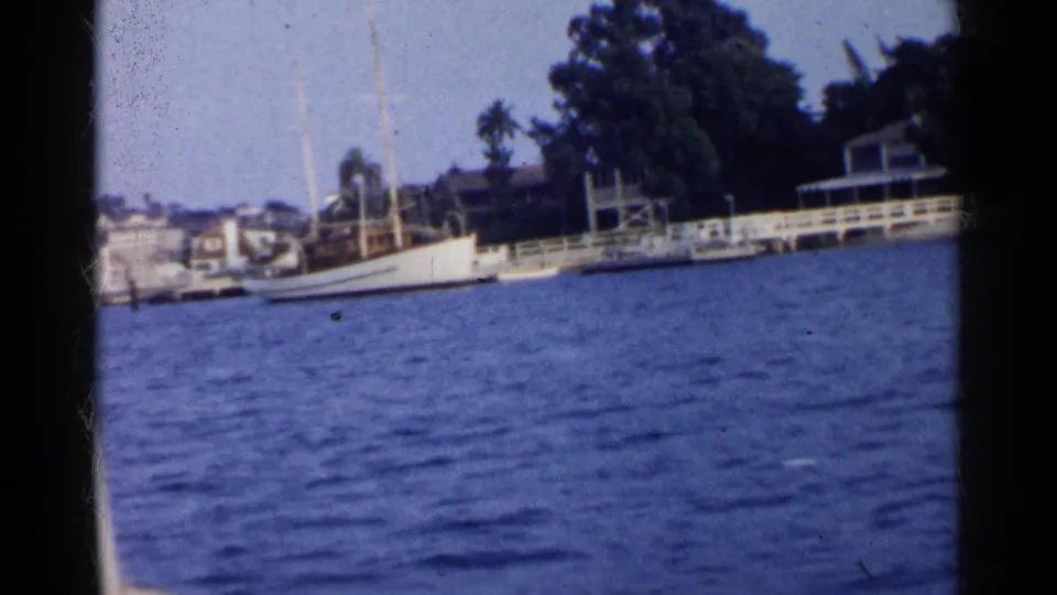 1955 California 8mm frame of a white sailboat moored by a dock, backed by shoreline homes and trees.