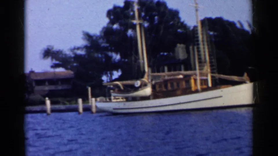 Close 1955 California waterfront frame of a white sailboat at dock posts, with trees and shoreline structures behind.