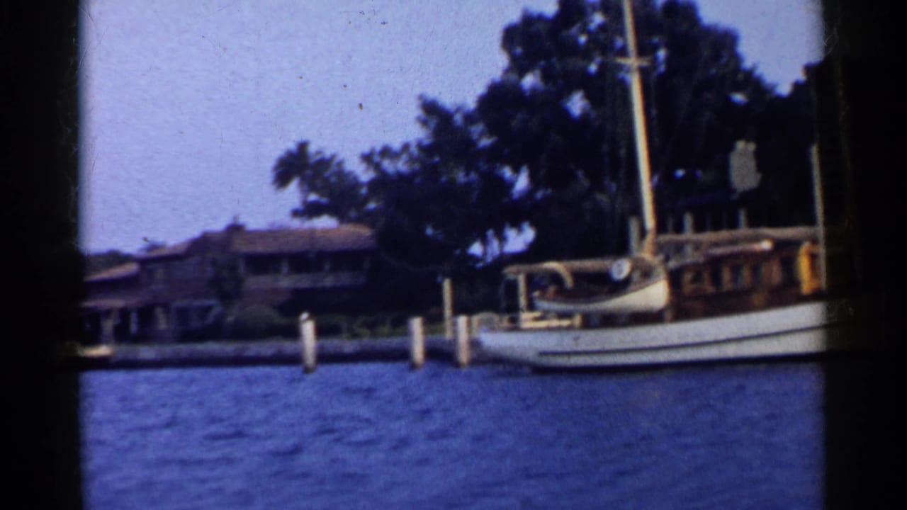 Sailboat docked beside a wooden pier with waterfront houses and trees behind it.