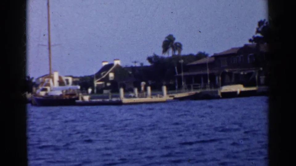 1955 8mm marina frame with a sailboat at left, white pier posts, waterfront homes, palms, and blue water.