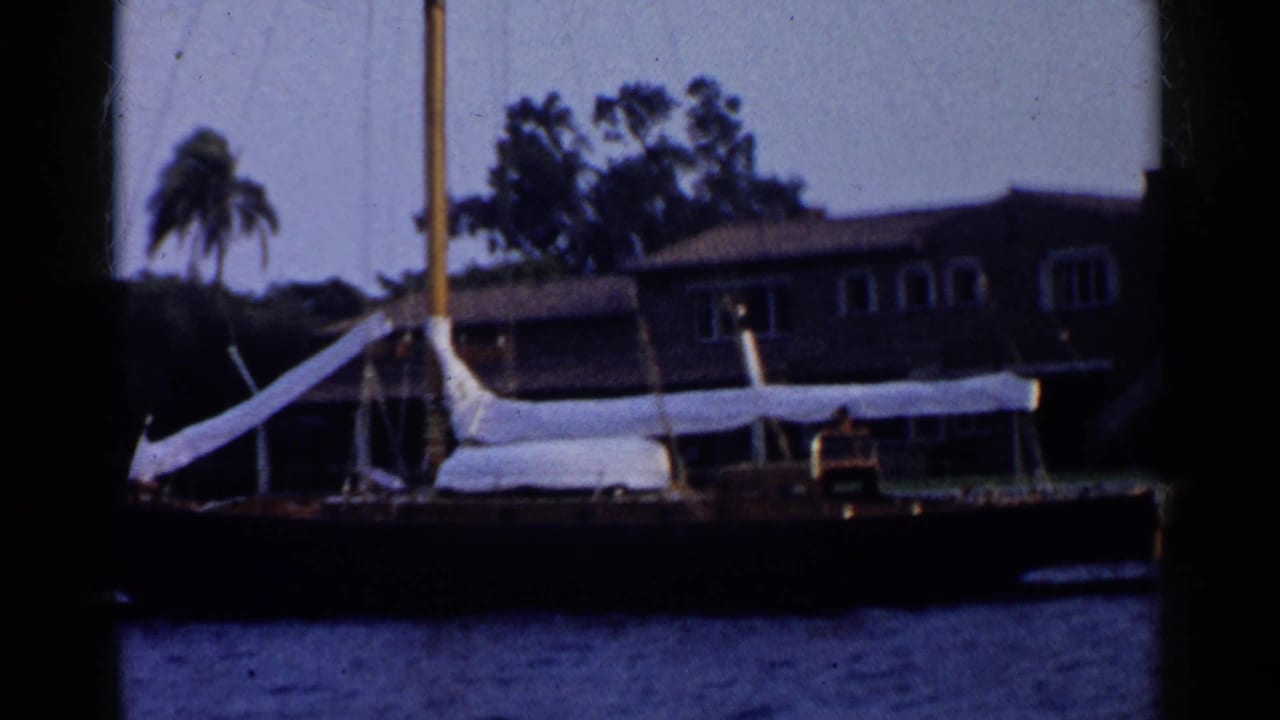 1955 California 8mm frame showing a sailboat at dock before a waterfront house, with a palm tree and dark film borders.