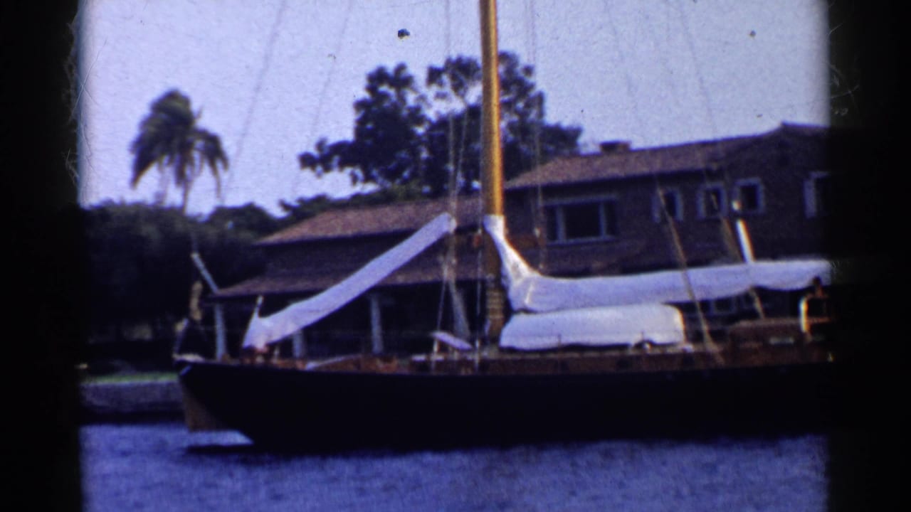 Closer view of a sailboat beside a waterfront house with trees behind it.