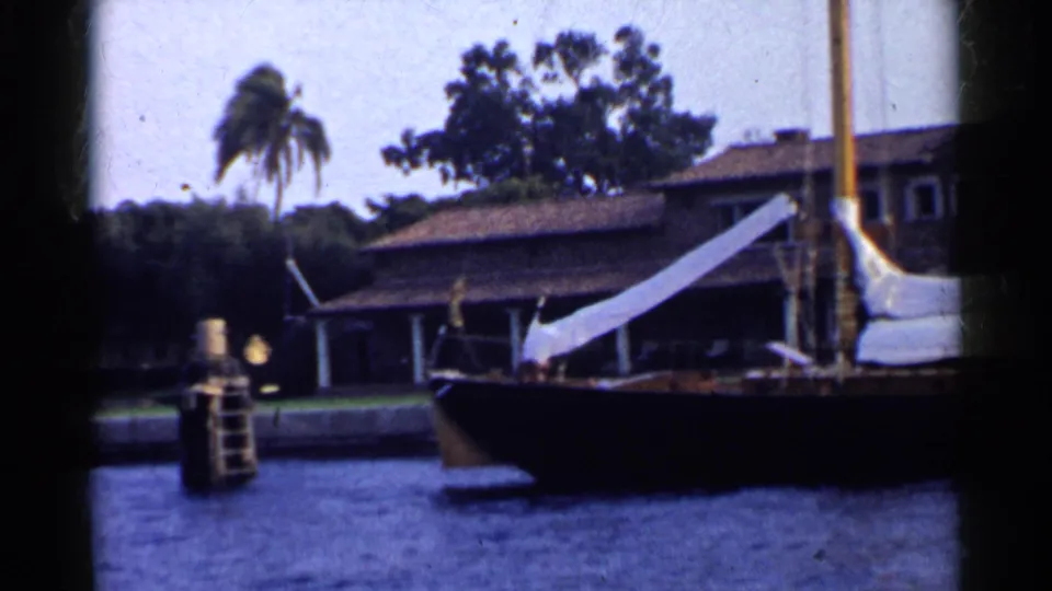 1955 California 8mm still showing the sailboat bow near a dock ladder, blue water, a low tiled-roof house, and a palm tree.
