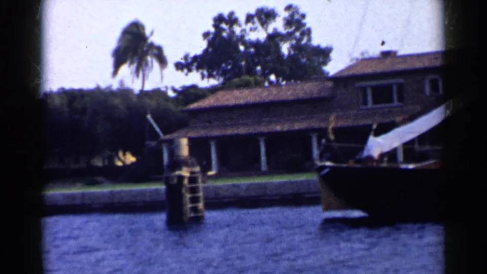 1955 California home-movie frame with a dock ladder, partial sailboat at right, blue water, tiled-roof house, palm tree, and soft 8mm color.