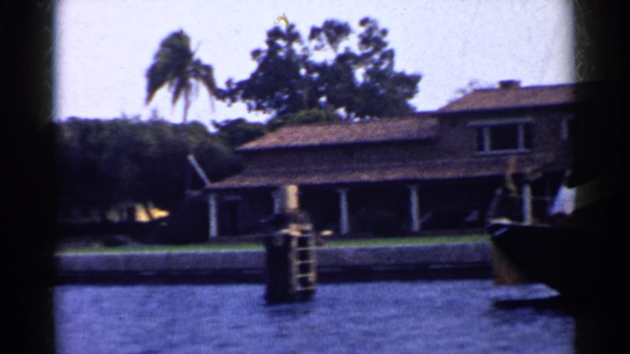 Waterfront stucco house with tiled roof, columns, lawn, palms, and dock post seen from the water.