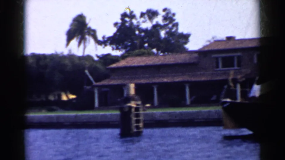 1955 California 8mm frame of a waterfront residence with tiled roof, white columns, palms, lawn, and a dock post in the foreground.
