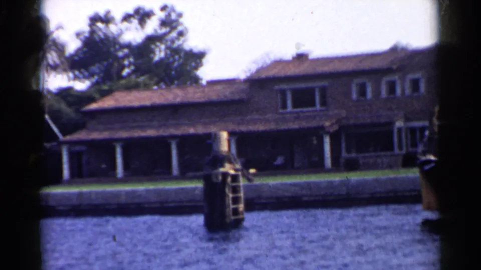 1955 California shoreline view from a boat, showing a large waterfront house, lawn, dock post with ladder, trees, and a partial boat edge.