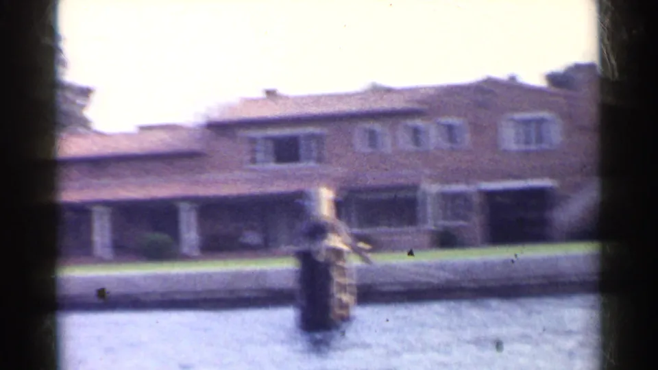 Soft 8mm frame from 1955 California showing a closer boat-level view of a waterfront house, lawn, columns, roofline, and dock post.