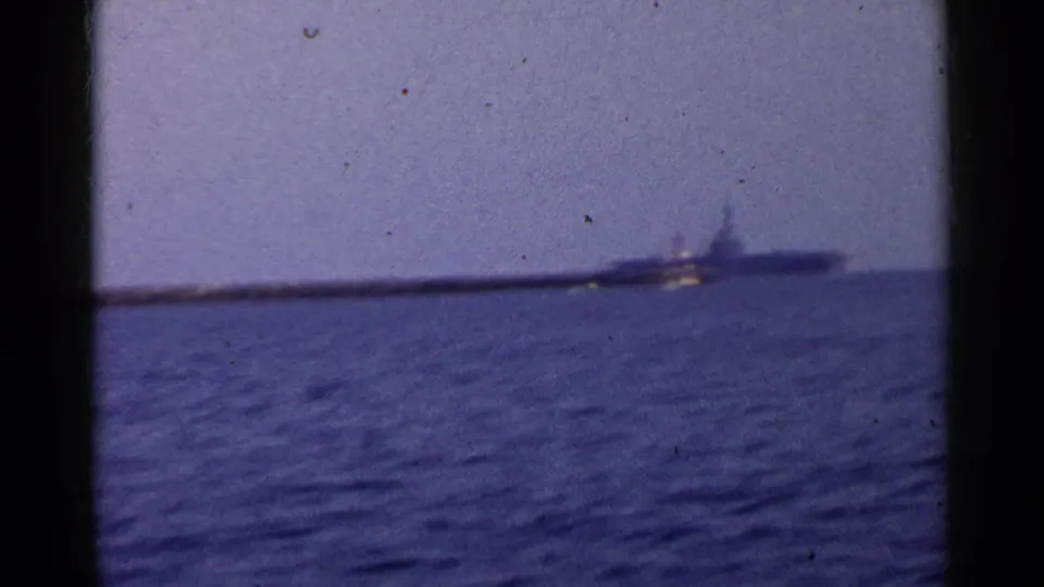 1955 California 8mm frame of open water with a distant breakwater or shoreline and a naval-looking ship on the horizon.