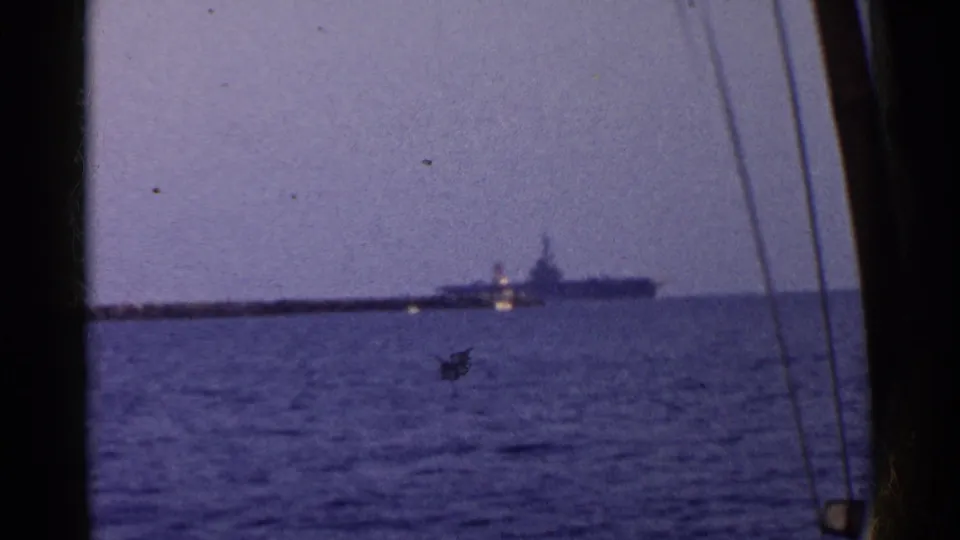 1955 California 8mm frame showing a distant large naval vessel on open water, seen through dark framing and possible rigging or window lines.