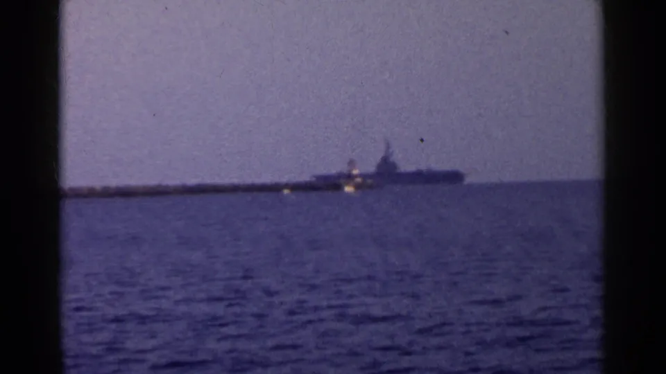 Distant large naval vessel, likely an aircraft carrier, moves along the horizon over blue open water in a faded 1955 California home-movie frame.