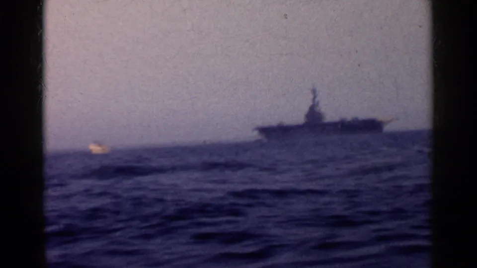 1955 California home-movie frame with a large naval vessel at right and a small bright boat or wake on the water nearby.