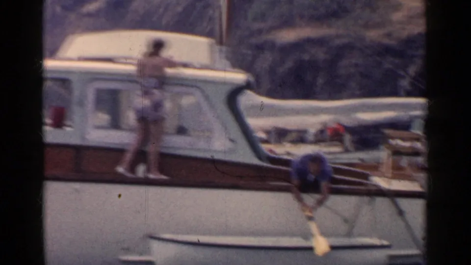 1955 California 8mm frame showing a woman in a swimsuit standing on a small cabin boat as another person paddles near the bow.