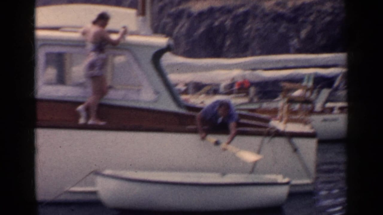 Two adults prepare a small rowboat beside a cabin cruiser on calm water.