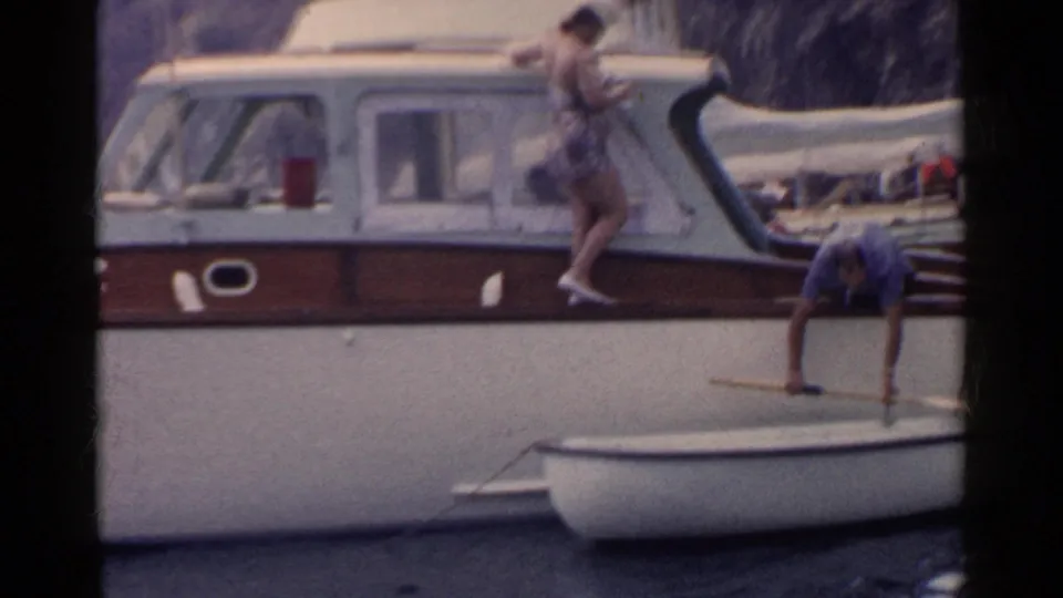 1955 California boating frame with a woman in a swimsuit on the cabin cruiser and a man reaching down toward the rowboat.