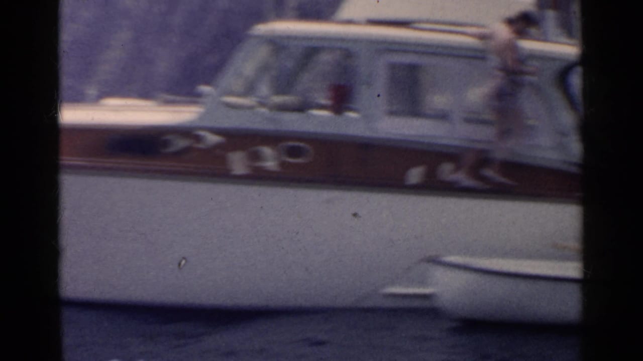 1955 California home-movie frame showing a white motorboat with brown trim, a small dinghy, and adults preparing near the water.
