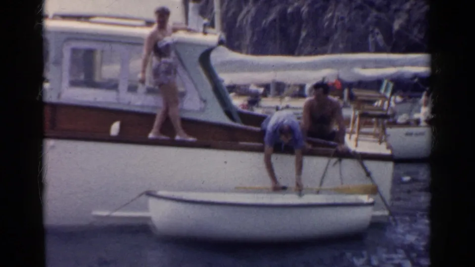 1955 California home-movie frame showing a woman on the motorboat deck and a man leaning into a dinghy near the water.