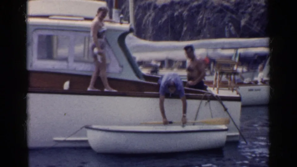1955 California home-movie frame showing adults around a small white rowboat beside a white motorboat with wooden trim.