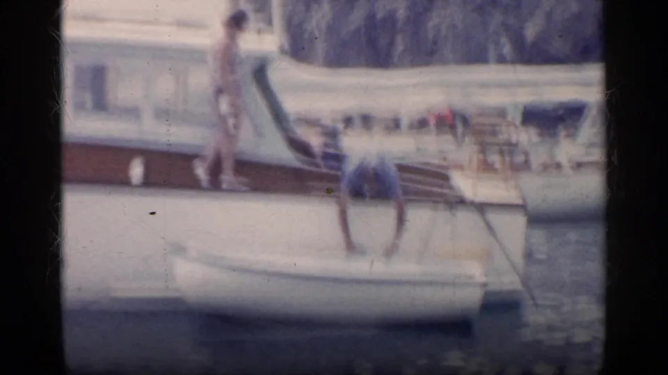 Soft 8mm frame from the California boating scene with a man leaning into a white rowboat near a motorboat and another adult standing on deck.