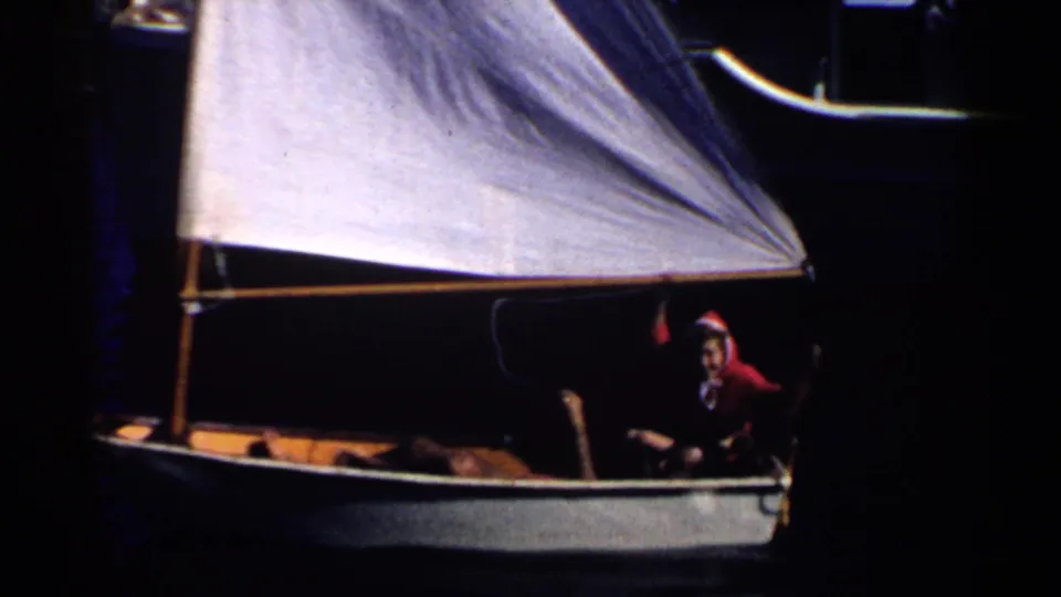 Nighttime 1955 California boating frame showing a small sailboat with an illuminated white sail and a seated person in a red hooded jacket.