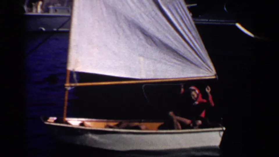 Low-light 8mm frame of a small sailboat under a white sail, with a person in a red hooded jacket steering or gesturing from the stern.