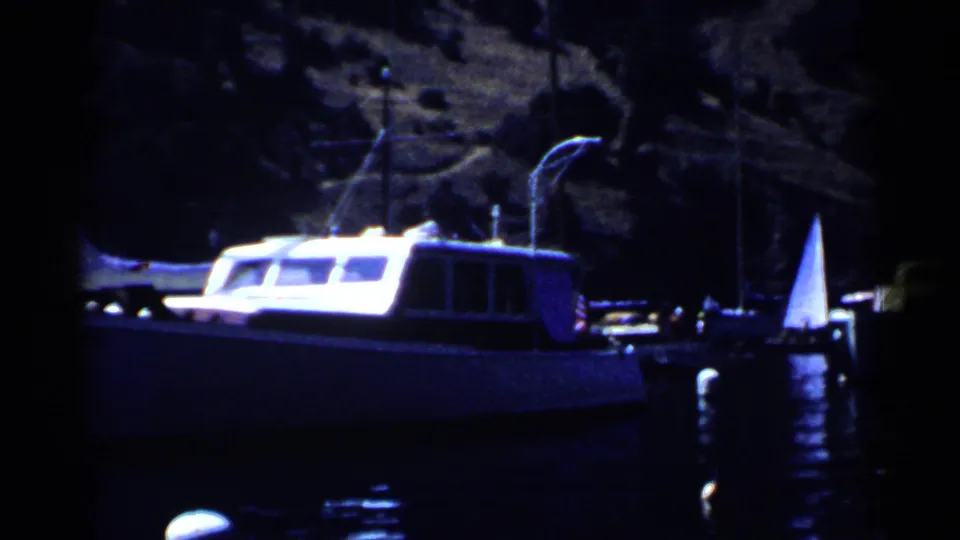 1955 California 8mm frame showing a cabin motorboat, a small sailboat, mooring posts, and a dark shoreline.
