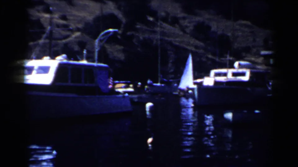 1955 California marina frame with moored small boats, a white sail, dark water, and rocky shoreline in soft 8mm color.