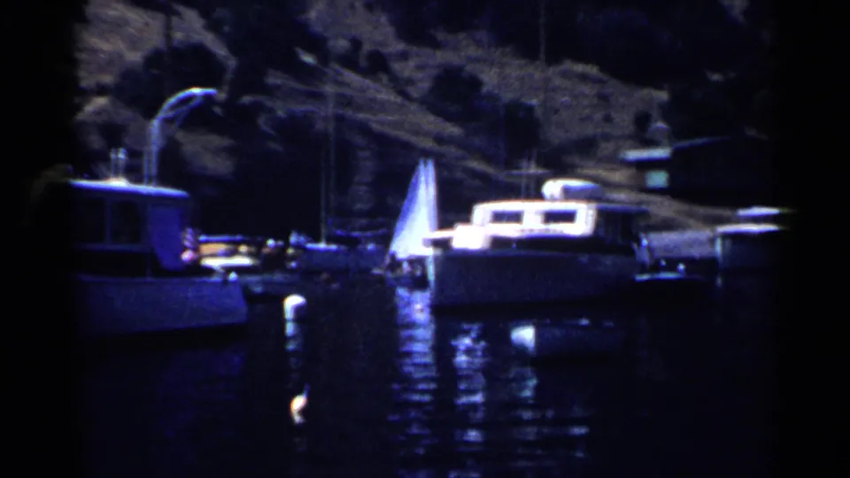 1955 California 8mm frame showing motorboats and a sailboat docked near a shadowed shoreline.