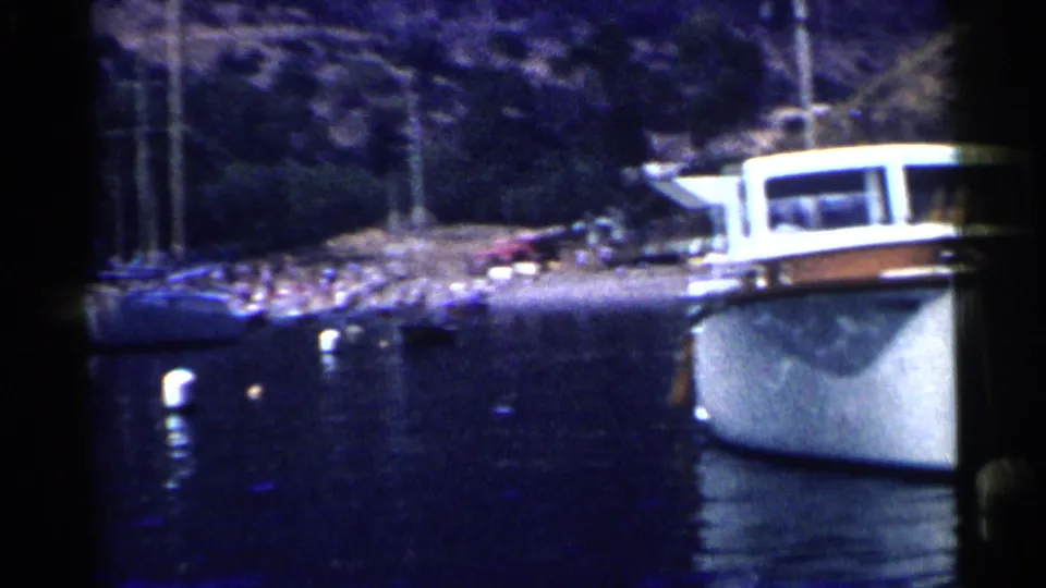 1955 California harbor frame with a large white motorboat, moored sailboats, and distant waterfront figures.