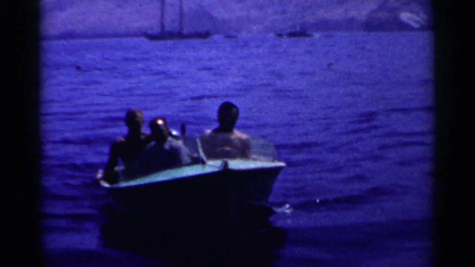 Three men in a small motorboat pass across blue coastal water, with distant sailboats and hazy mountains in the background.