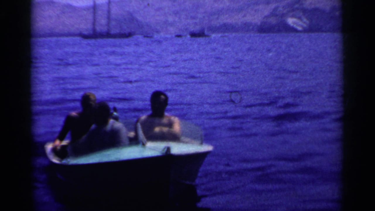 California 1955 home-movie frame showing three adult men in a small boat against blue lake water.