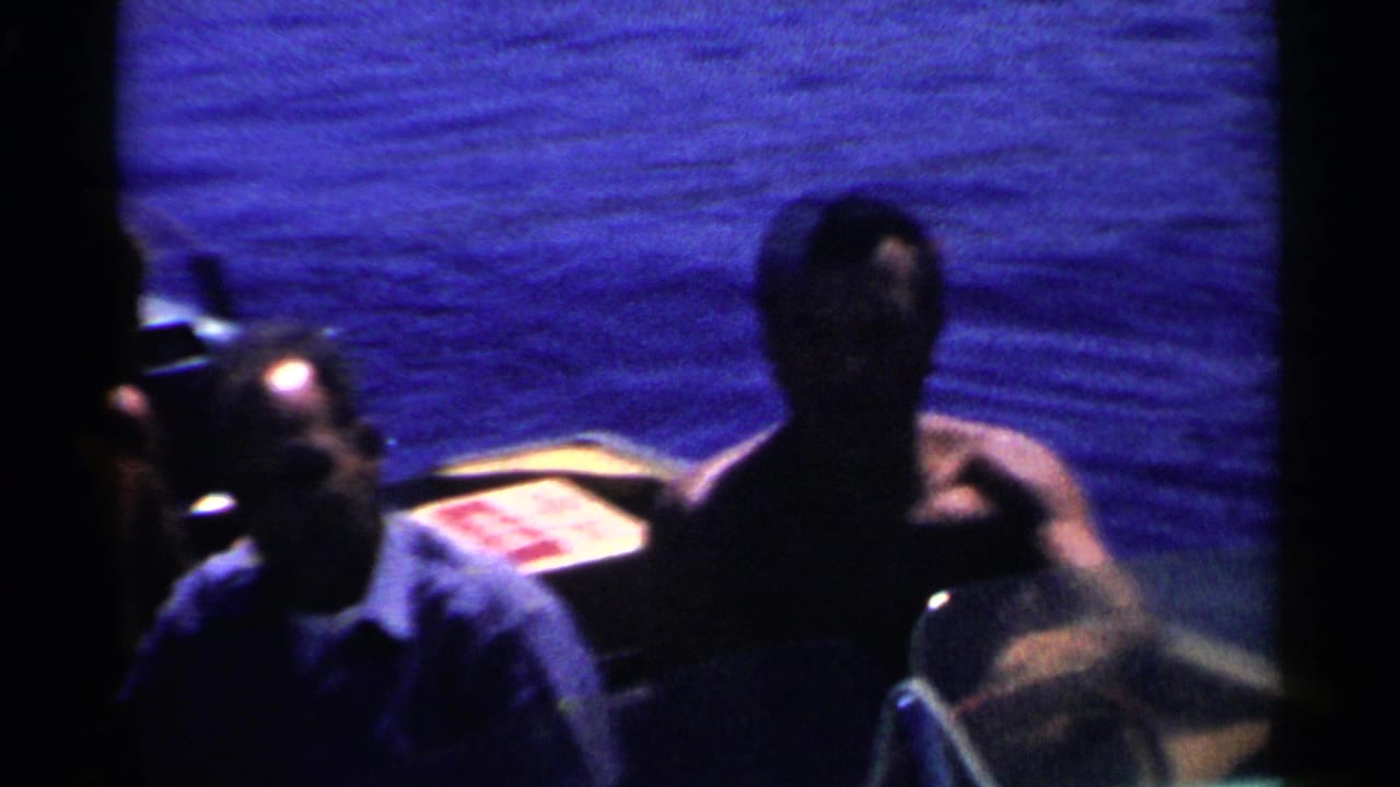 Shadowed close view of two adult men by a boat, from a 1955 California 8mm lake outing.