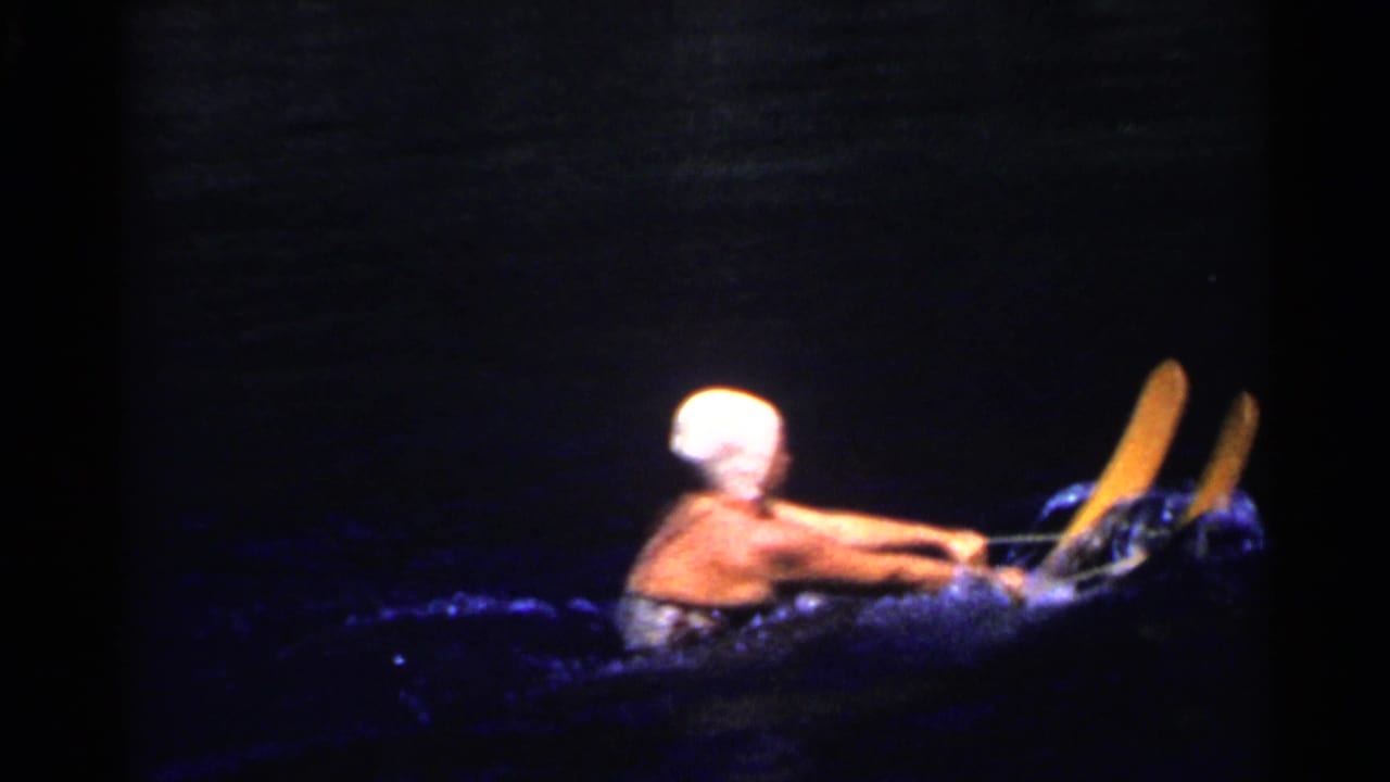 1955 California home-movie frame of a water skier wearing a white cap and riding yellow skis across dark water.